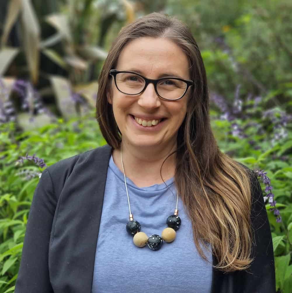 Nicole Kopel is a Dietitian in Melbourne assisting people with Eating Disorders and concerns with body image, weight, and eating behaviours. She is wearing a purple top with a beaded necklace. She has long brown hair and glasses and is smiling at the camera.